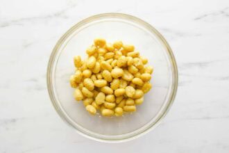 A clear glass bowl filled with seasoned Sheet Pan Gnocchi rests on a white marble surface.
