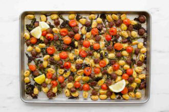 A sheet pan gnocchi dinner with roasted cherry tomatoes, kale, sausage slices, and lemon wedges arranged on top.