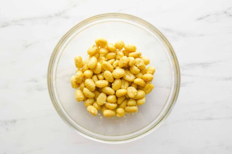 A clear glass bowl filled with seasoned Sheet Pan Gnocchi rests on a white marble surface.