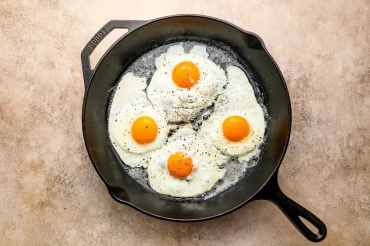 Four sunny-side-up eggs sizzle in a black cast iron skillet, seasoned with cracked black pepper, on a light brown countertop—perfect for a classic steak and eggs breakfast.