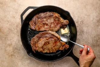 Two cooked steaks sizzle in a cast-iron skillet with a pat of melting butter; a hand holds a spoon, ready to baste the steak to perfection.