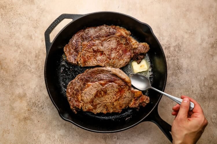Two cooked steaks sizzle in a cast-iron skillet with a pat of melting butter; a hand holds a spoon, ready to baste the steak to perfection.