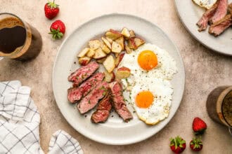 A plate of steak and eggs with roasted potatoes is served alongside strawberries and cups of coffee, all arranged on a light-colored surface.