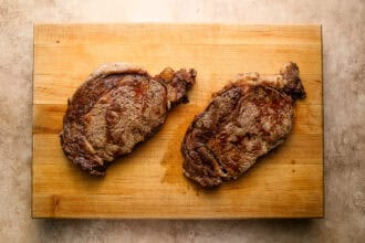 Two cooked ribeye steaks rest on a wooden cutting board, with a light brown textured surface underneath—perfect for a classic steak and eggs breakfast.