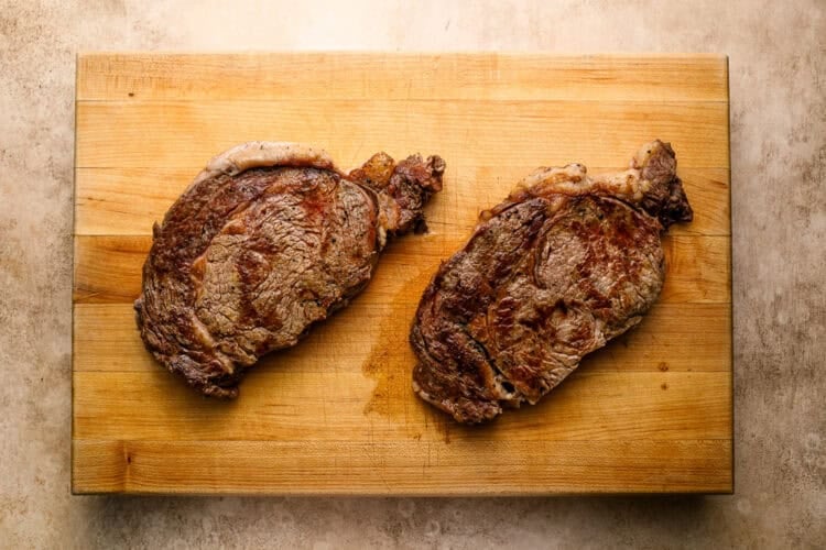 Two cooked ribeye steaks rest on a wooden cutting board, with a light brown textured surface underneath—perfect for a classic steak and eggs breakfast.