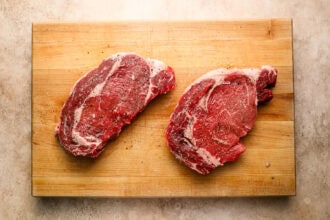 Two raw, seasoned ribeye steaks on a wooden cutting board placed on a light, textured surface—perfect for your next steak and eggs breakfast.