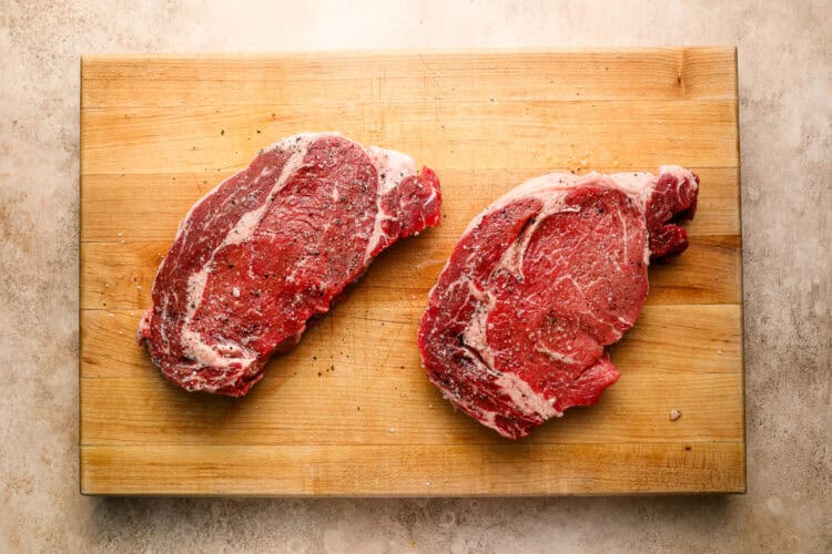 Two raw, seasoned ribeye steaks on a wooden cutting board placed on a light, textured surface—perfect for your next steak and eggs breakfast.