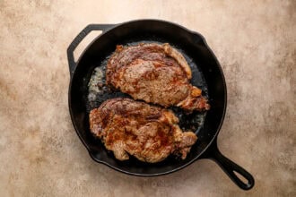 Two cooked ribeye steaks in a black cast iron skillet on a light brown textured surface, perfect for a hearty Steak and Eggs breakfast.