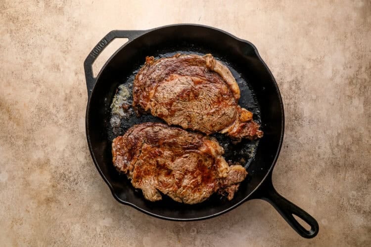 Two cooked ribeye steaks in a black cast iron skillet on a light brown textured surface, perfect for a hearty Steak and Eggs breakfast.