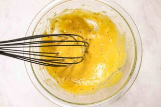 A whisk mixing yellow vinaigrette with black pepper and spices in a clear glass bowl on a light-colored surface, perfect as a zesty Pourhouse Caesar dressing.