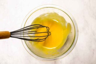 A metal whisk with a wooden handle rests in a glass bowl containing a yellow mixture on a white surface, ready to mix the perfect Pourhouse Caesar cocktail.