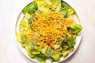 A plate of chopped romaine lettuce topped with shredded cheddar, grated parmesan, and a hint of Pourhouse Caesar on a white surface.