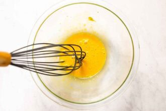 A metal whisk and beaten egg yolks in a clear glass bowl on a white surface, ready to craft the perfect Caesar cocktail.