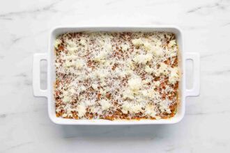 A white baking dish filled with zucchini lasagna, topped with shredded cheese and dollops of ricotta, sits on a white marble surface.