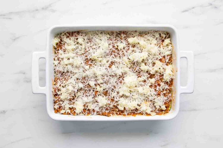 A white baking dish filled with zucchini lasagna, topped with shredded cheese and dollops of ricotta, sits on a white marble surface.