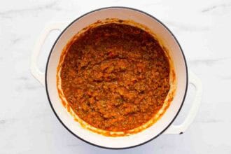 A white pot filled with cooked meat and tomato sauce, sits on a white marble surface.