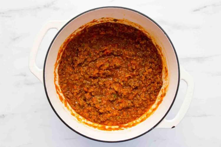 A white pot filled with cooked meat and tomato sauce, sits on a white marble surface.