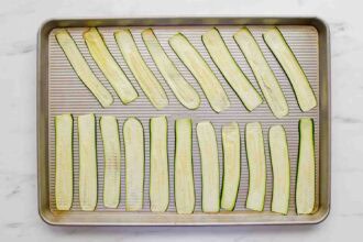 A baking sheet with twenty evenly spaced, raw zucchini slices arranged in rows on a white surface.