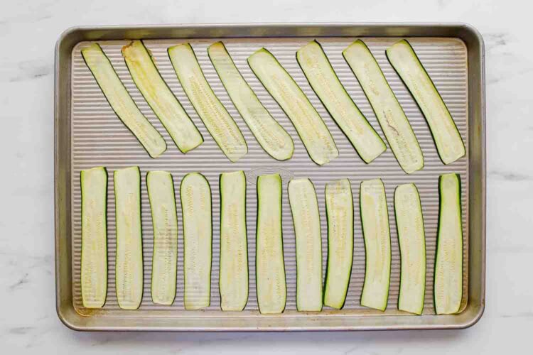 A baking sheet with twenty evenly spaced, raw zucchini slices arranged in rows on a white surface.