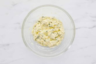 A clear glass bowl on a marble surface contains a mixture of ricotta cheese, herbs, and grated cheese.