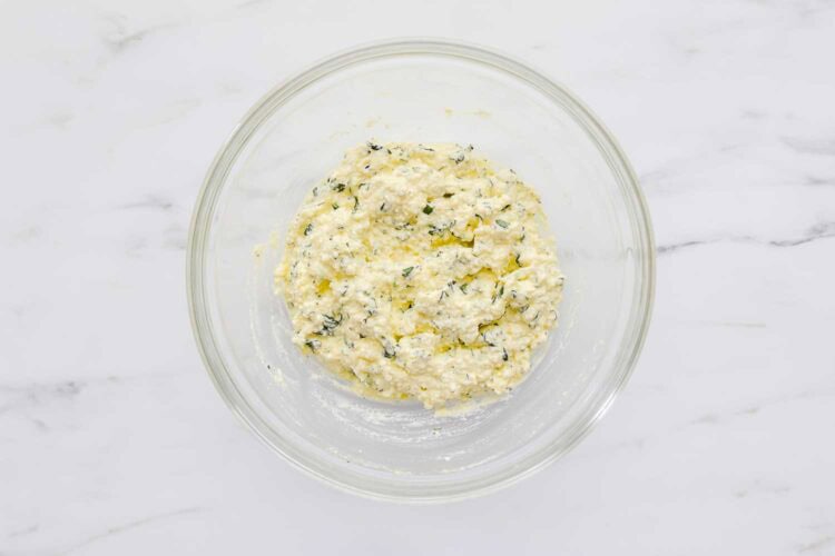 A clear glass bowl on a marble surface contains a mixture of ricotta cheese, herbs, and grated cheese.