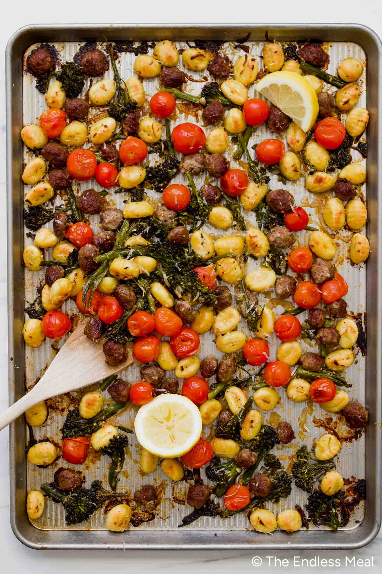 Sheet Pan Gnocchi with roasted cherry tomatoes, broccolini, sausage pieces, and lemon wedges, all deliciously caramelized on one pan—a wooden spoon rests nearby for easy serving.