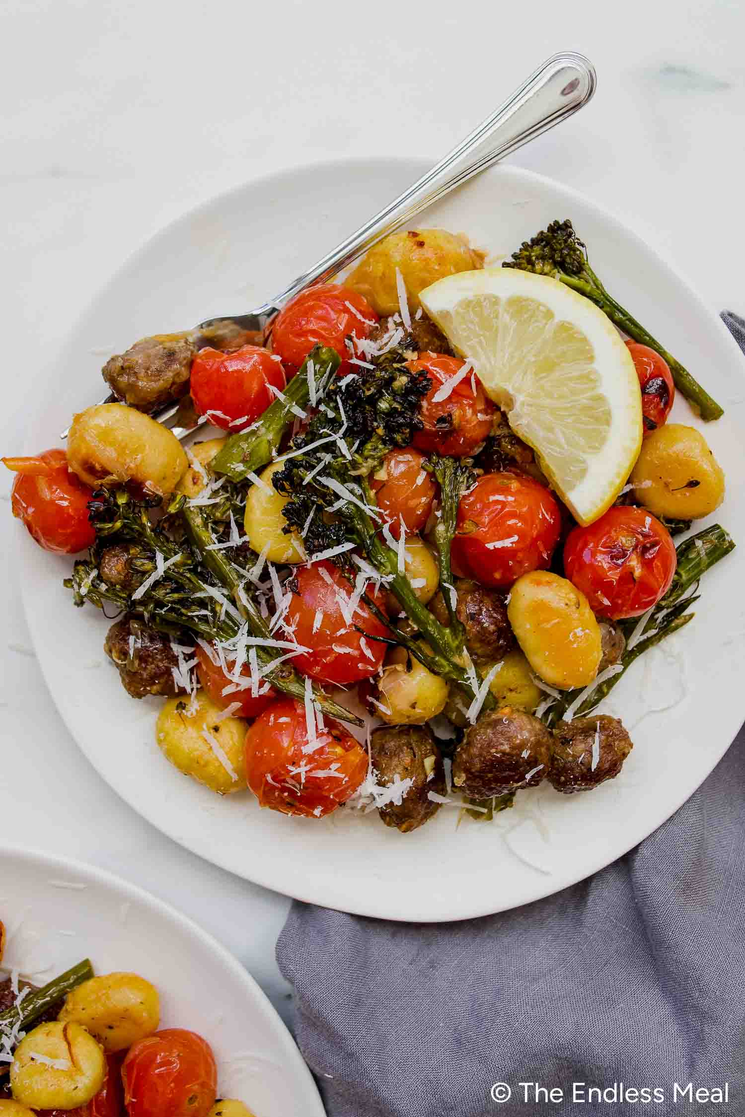 A plate of Sheet Pan Gnocchi with roasted cherry tomatoes, broccolini, meatballs, grated cheese, and a lemon wedge, served with a fork.
