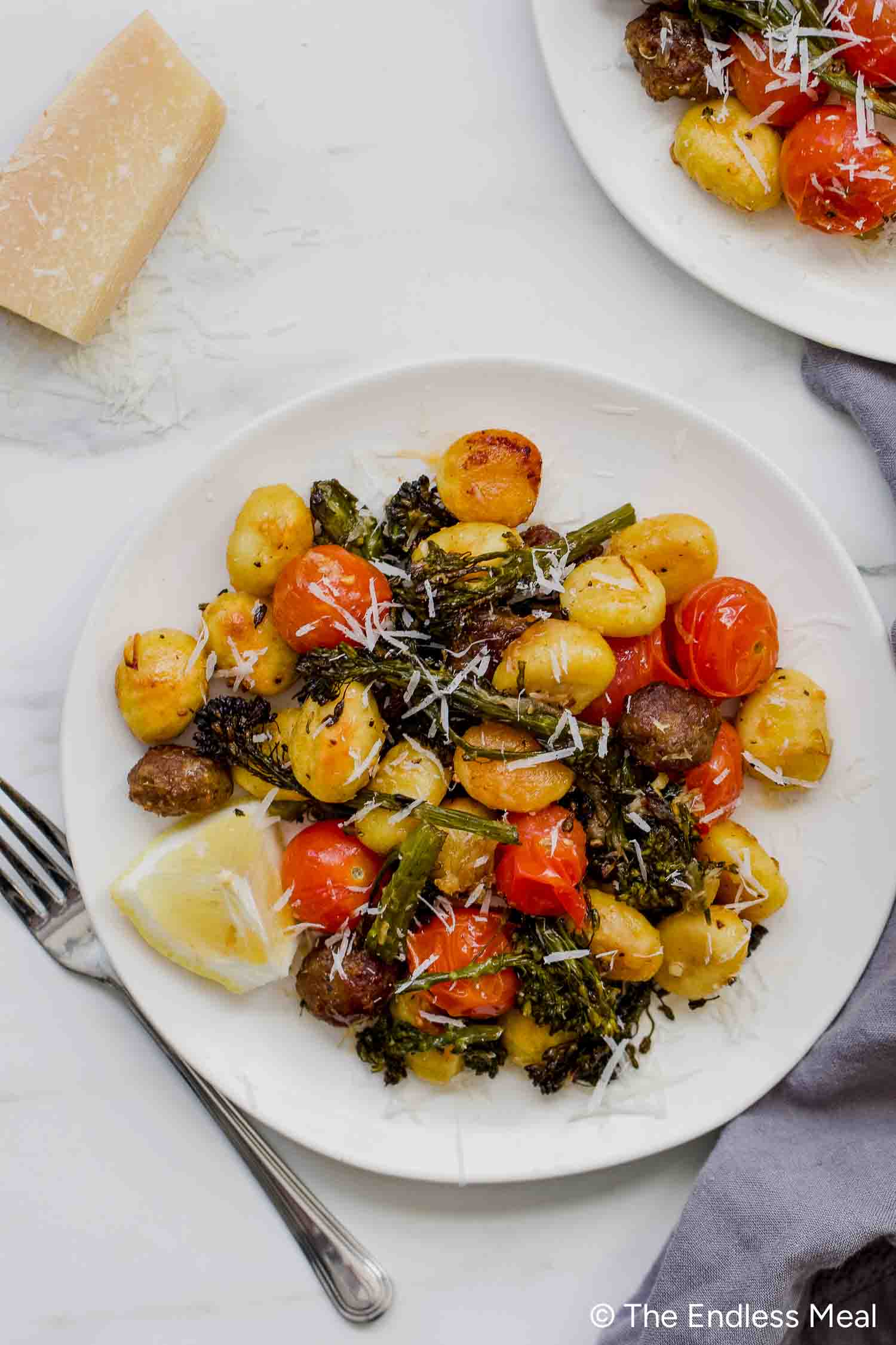 A white plate with Sheet Pan Gnocchi, roasted cherry tomatoes, broccolini, sausage pieces, and grated cheese, served with a lemon wedge on the side.