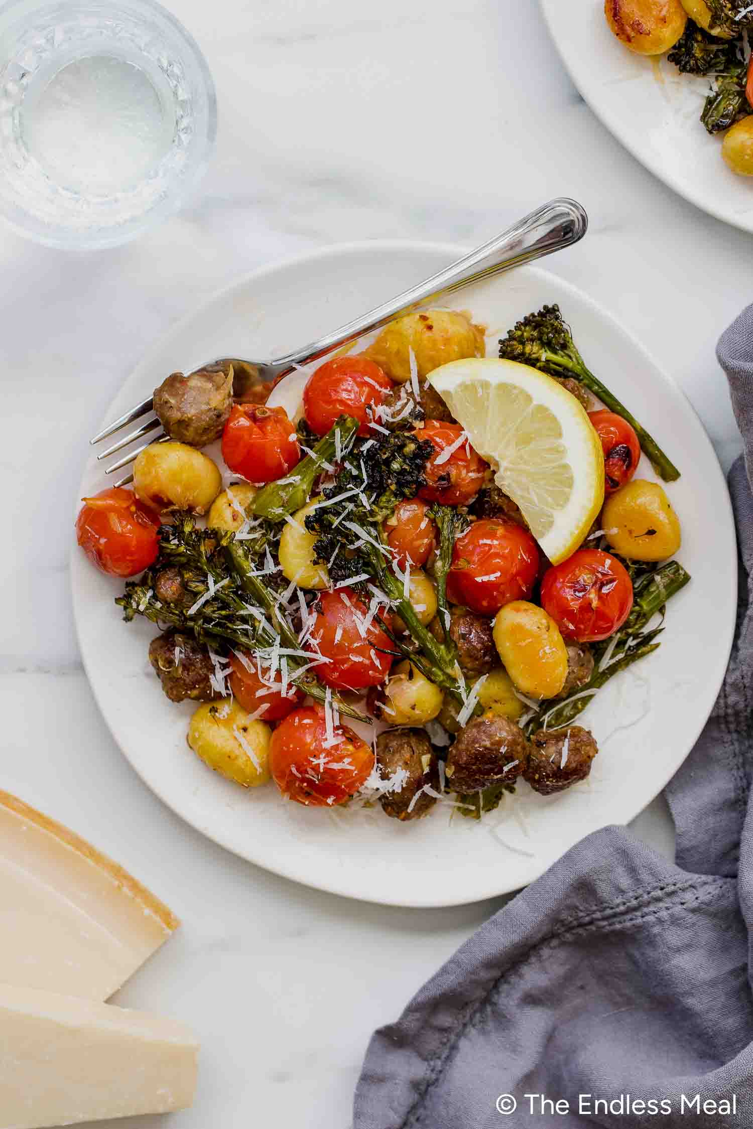 A plate of Sheet Pan Gnocchi with roasted cherry tomatoes, broccolini, mushrooms, grated cheese, and a lemon wedge, served with a fork.