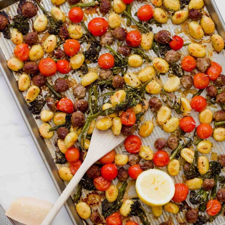 A Sheet Pan Gnocchi dinner featuring roasted gnocchi, cherry tomatoes, broccolini, sausage slices, and lemon halves, all served with a wooden spoon on top.