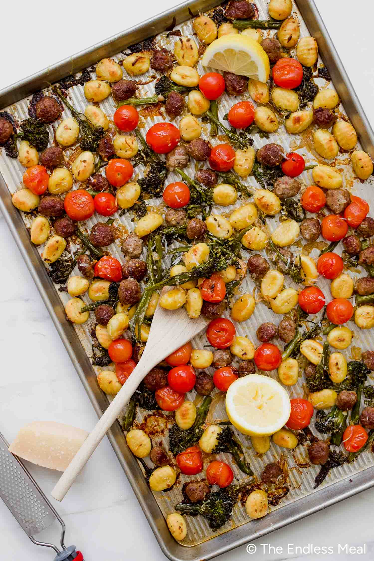 A Sheet Pan Gnocchi dinner featuring roasted gnocchi, cherry tomatoes, broccolini, sausage slices, and lemon halves, all served with a wooden spoon on top.