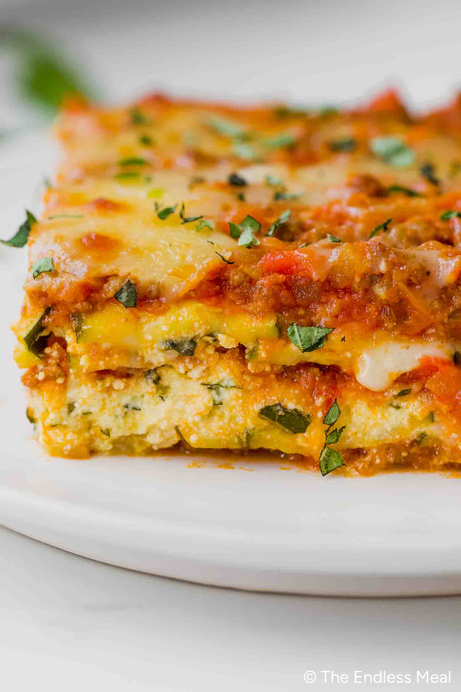A close-up of a slice of zucchini lasagna with layers of pasta, cheese, tomato sauce, and herbs, served on a white plate.