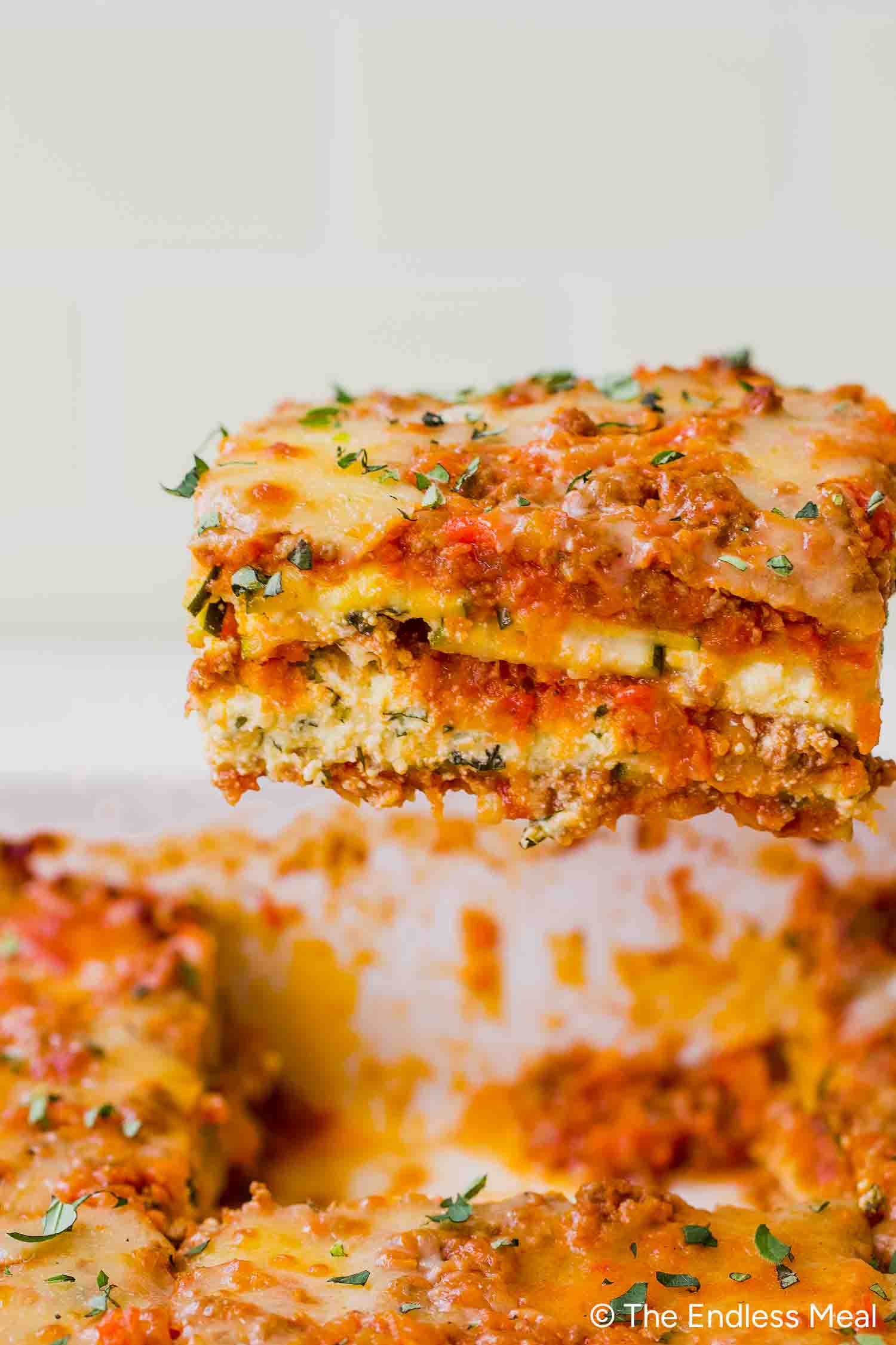 A slice of zucchini lasagna with layers of pasta, meat sauce, ricotta cheese, and herbs is being lifted from a baking dish.