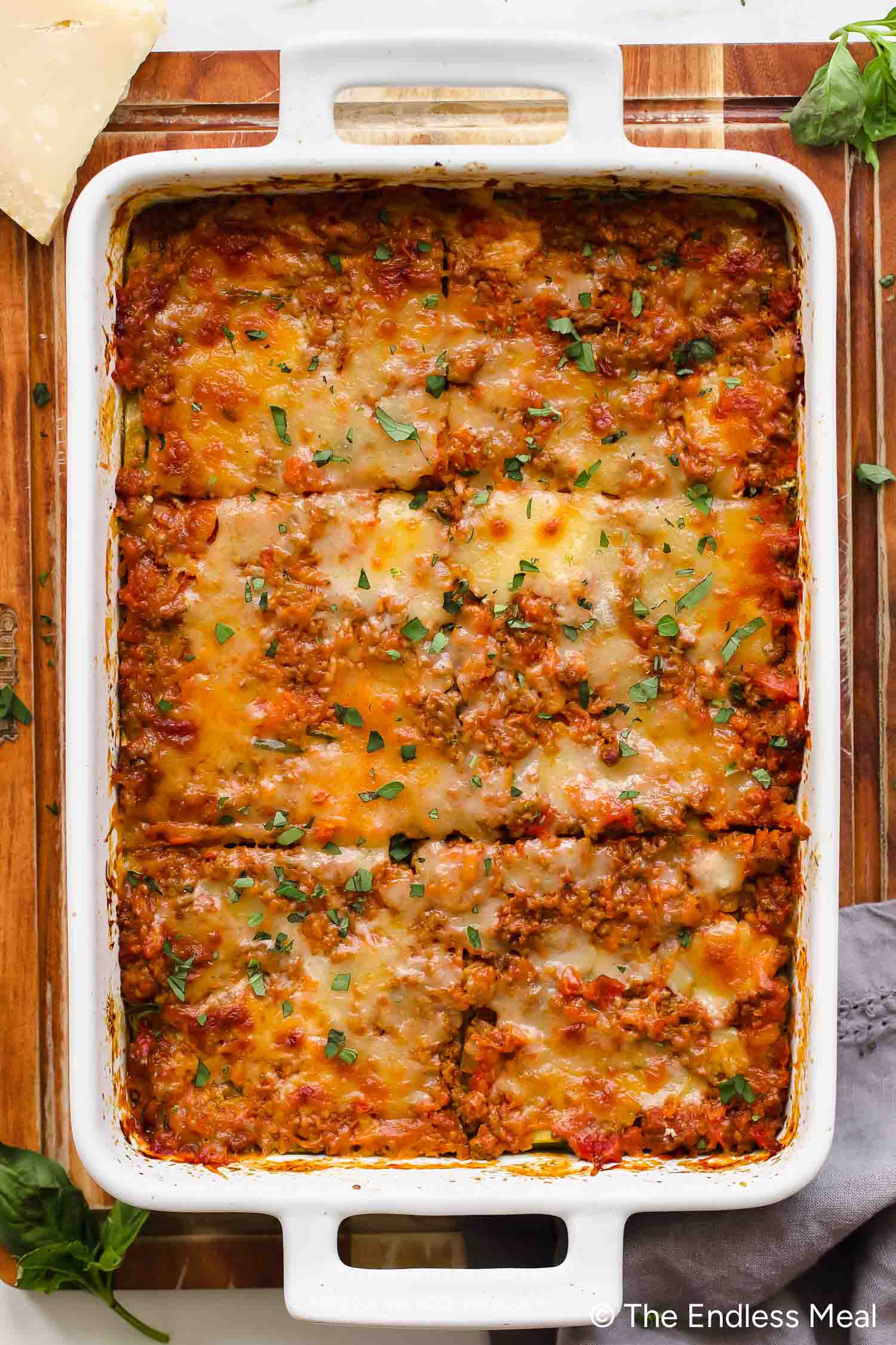 A baked zucchini lasagna in a white rectangular dish, topped with melted cheese and garnished with chopped herbs on a wooden surface.