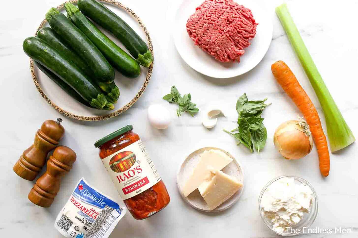 Ingredients for a Zucchini Lasagna recipe displayed on a white surface, featuring zucchini, ground beef, sauces, cheese, vegetables, and seasonings.