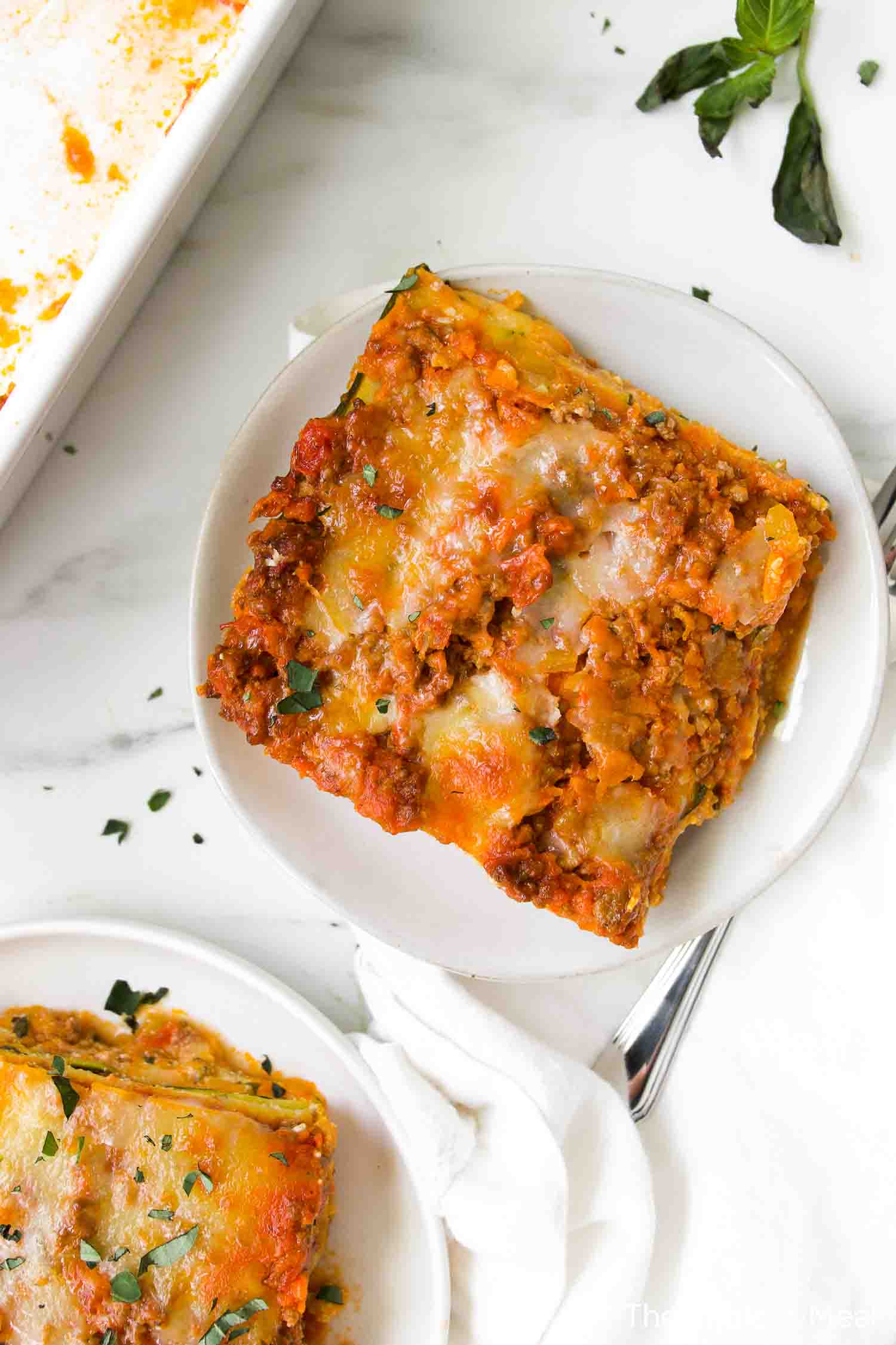 A square slice of baked zucchini lasagna with meat sauce and melted cheese on a white plate, garnished with chopped herbs.