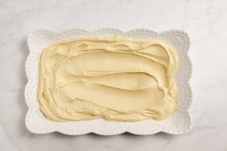 A rectangular white plate with scalloped edges holds a smooth, even layer of party-worthy hummus, perfect for adding your favorite hummus toppings, all displayed on a white marble surface.