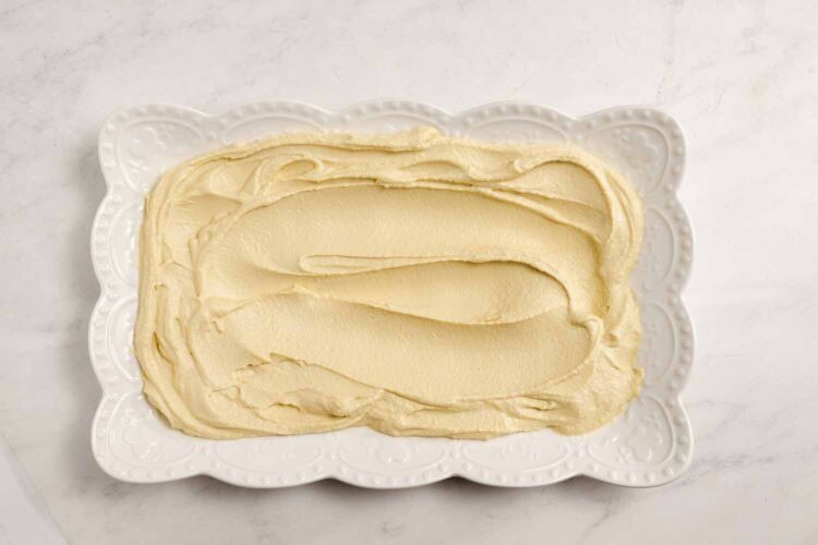 A rectangular white plate with scalloped edges holds a smooth, even layer of party-worthy hummus, perfect for adding your favorite hummus toppings, all displayed on a white marble surface.