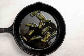 A black cast iron skillet with oil and several sage leaves frying inside—perfect for preparing unique hummus toppings—sits on a white surface.