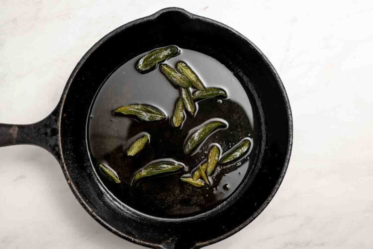 A black cast iron skillet with oil and several sage leaves frying inside—perfect for preparing unique hummus toppings—sits on a white surface.