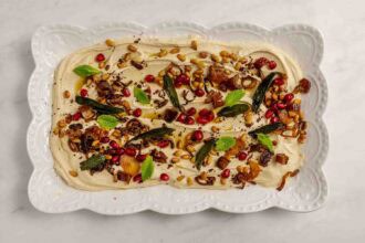 A rectangular white platter with party-worthy hummus, topped with pomegranate seeds, pine nuts, sautéed onions, dates, fried sage leaves, and fresh mint—an irresistible array of hummus toppings.
