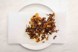 Crispy fried shallots and garlic, perfect as party-worthy hummus toppings, are draining on a paper towel atop a white plate.
