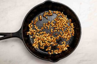 Pine nuts being toasted in a black cast iron skillet on a white marble surface—perfect as party-worthy hummus toppings.