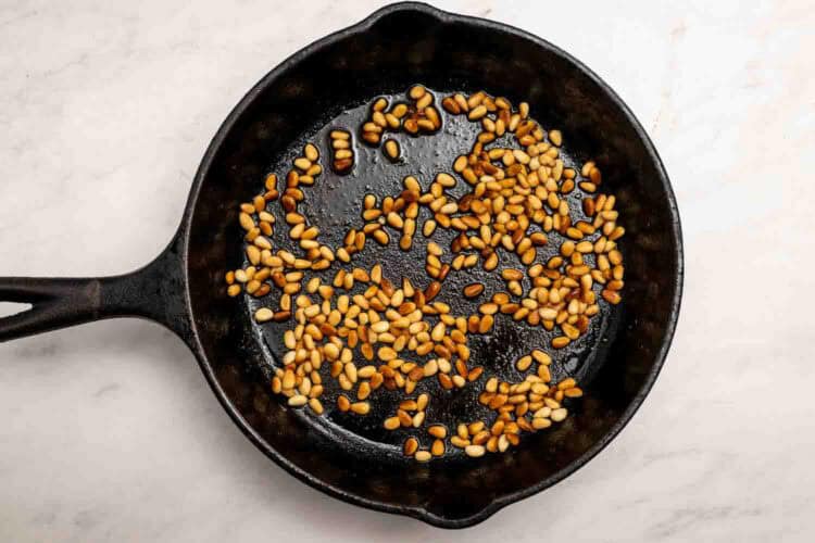 Pine nuts being toasted in a black cast iron skillet on a white marble surface—perfect as party-worthy hummus toppings.
