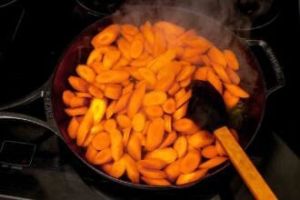 Sliced carrots being cooked in a black cast iron skillet with a wooden spatula, producing visible steam—perfect for making sticky carrots.