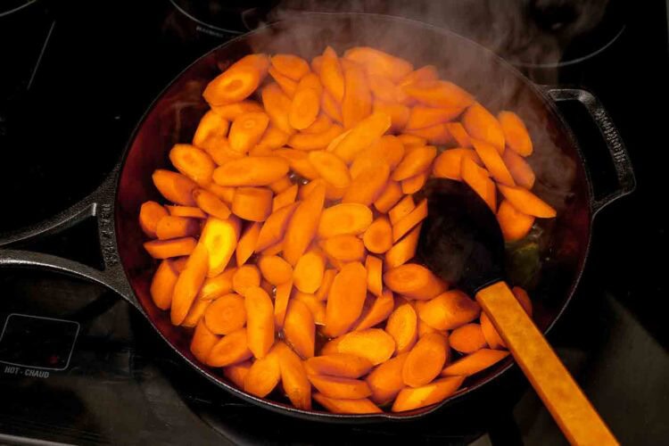 Sliced carrots being cooked in a black cast iron skillet with a wooden spatula, producing visible steam—perfect for making sticky carrots.