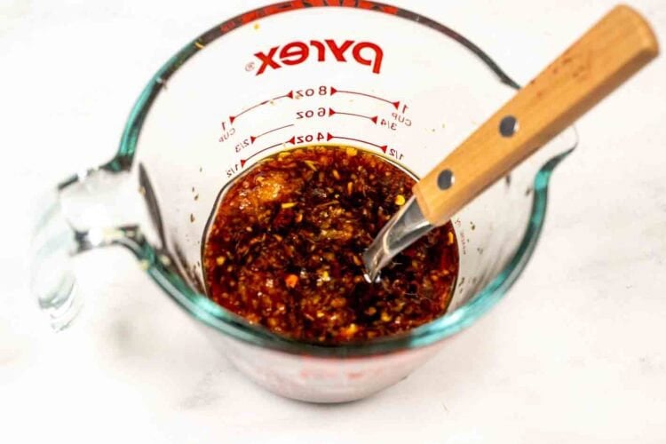 A glass Pyrex measuring cup filled with a chunky, reddish-brown sauce mixture for Sticky Carrots and a wooden-handled spoon on a white surface.