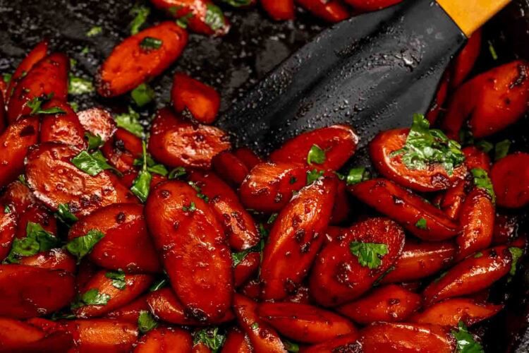 Sticky ginger soy glazed carrots garnished with chopped parsley in a pan, with a black spatula beside them.