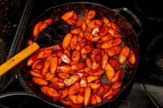 Ginger soy glazed carrots are sautéed in a cast iron skillet with spices, stirred with a black spatula on the stovetop for a flavorful side.