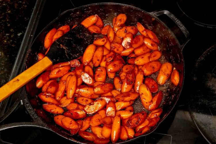 Ginger soy glazed carrots are sautéed in a cast iron skillet with spices, stirred with a black spatula on the stovetop for a flavorful side.